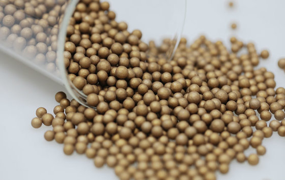 Gold-coloured alloy–ceramic composite balls spilling from a glass beaker (media maker's photo)